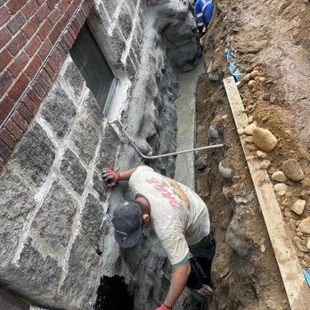 Worker applying cement to a stone foundation wall. Trench, brick building, and blue hard hat are visible.