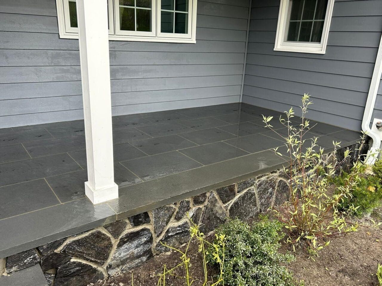 Gray tiled porch with white column and stone foundation, next to house with windows and greenery.