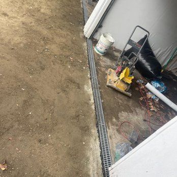 A trench drain alongside a building. Tools and debris surround the drain in a dirt area.