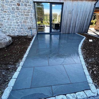 Outdoor patio with blue-gray stone tiles, a curved border, and a door leading to a building with stone and wood siding.