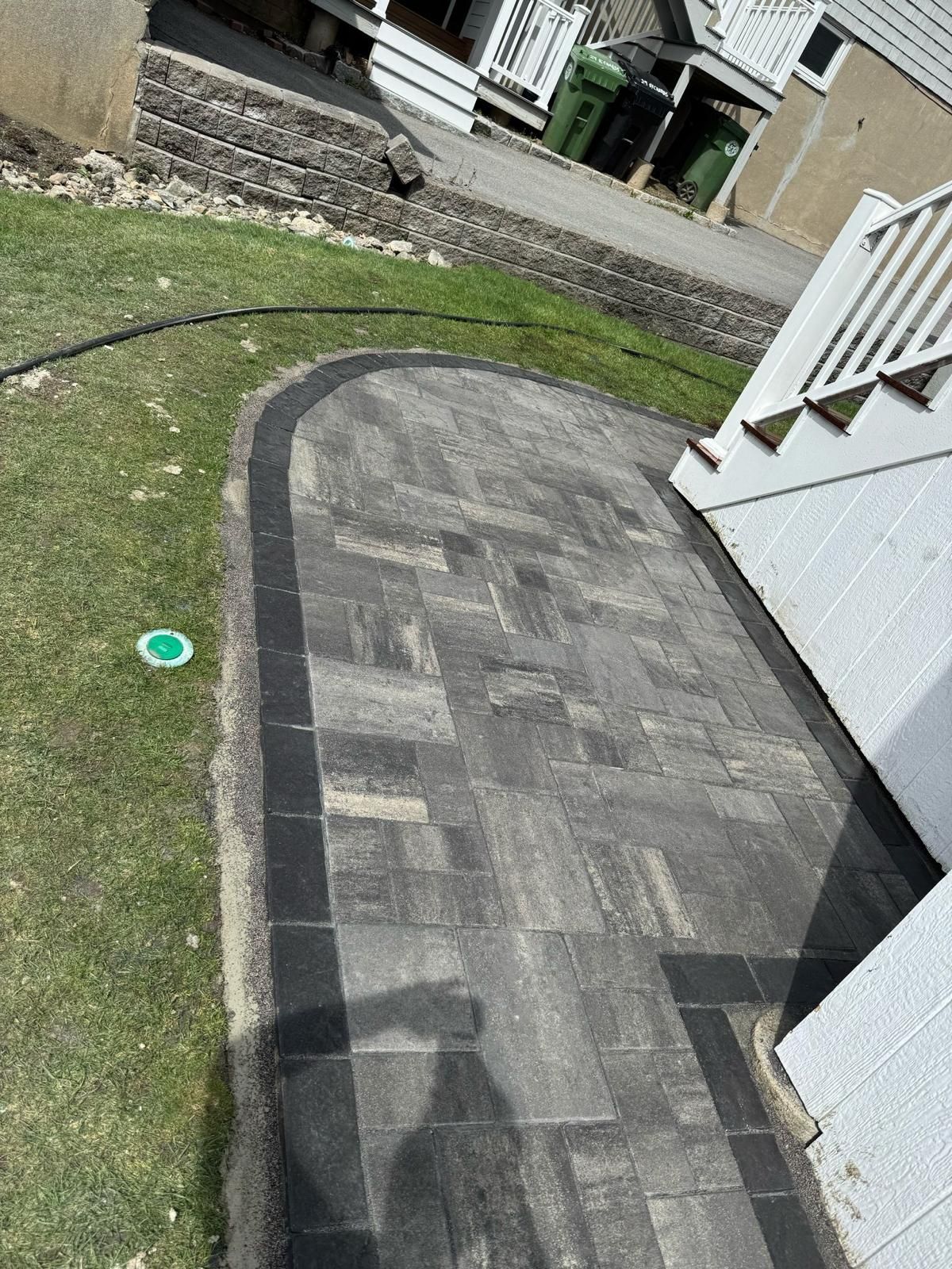 Patio with dark gray pavers, surrounded by grass and a black border. White stairs on the right.