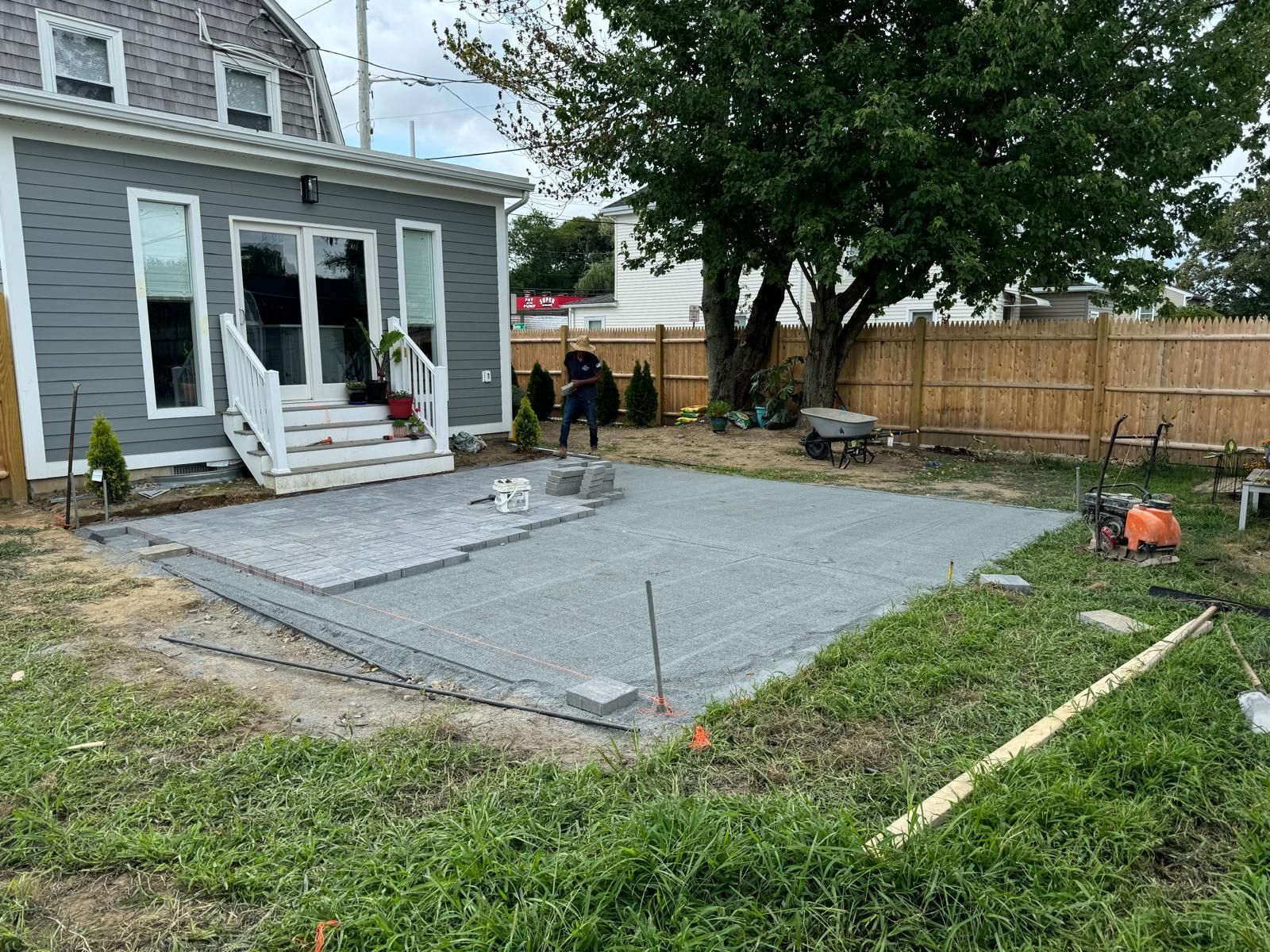 Backyard patio construction: gray gravel base, workers, house with glass doors, wooden fence.