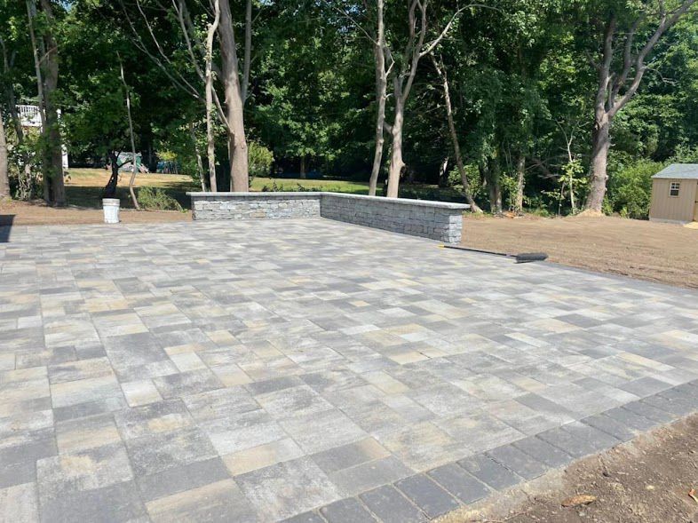 Paved patio with gray and beige rectangular pavers, a stone wall, and trees in the background.