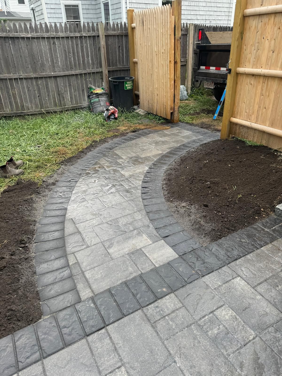 Brick pathway curves through a backyard, bordered by dark bricks and dirt. Wooden fences and grass surround it.
