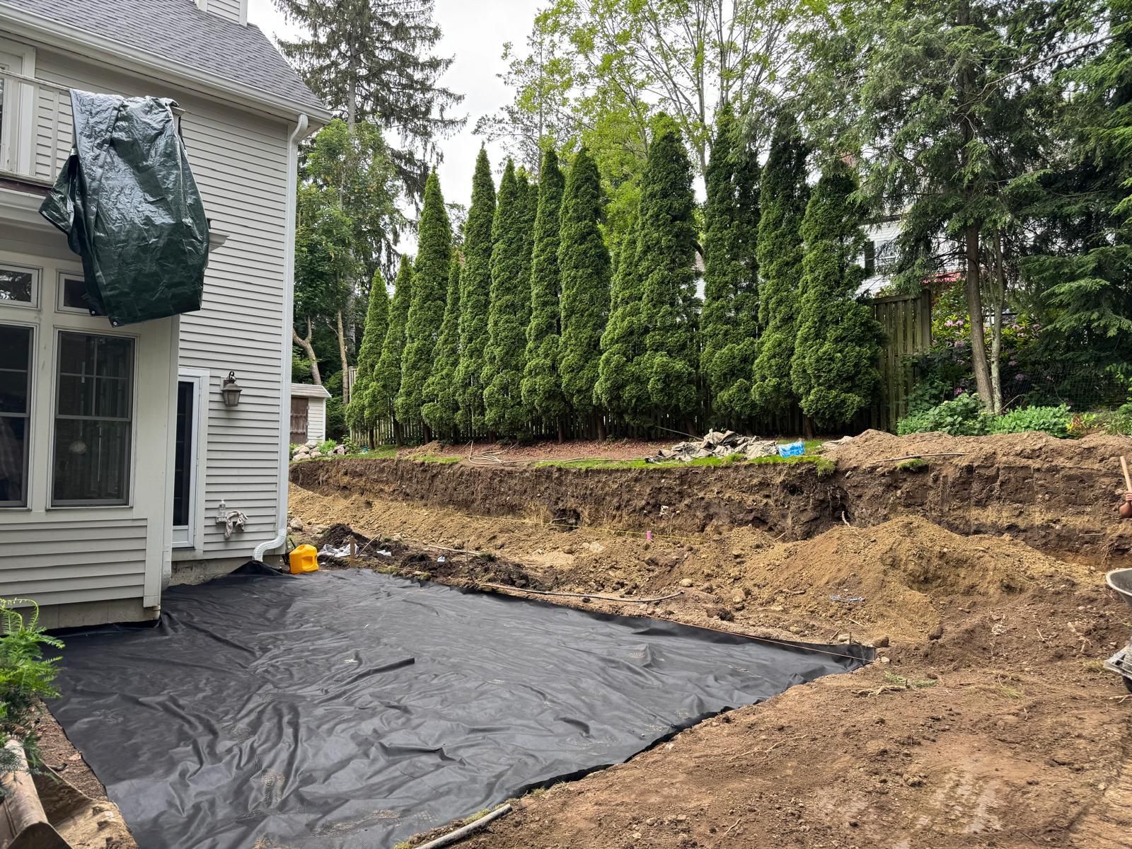 Backyard excavation for a patio; black fabric laid down; row of tall green trees in the background.