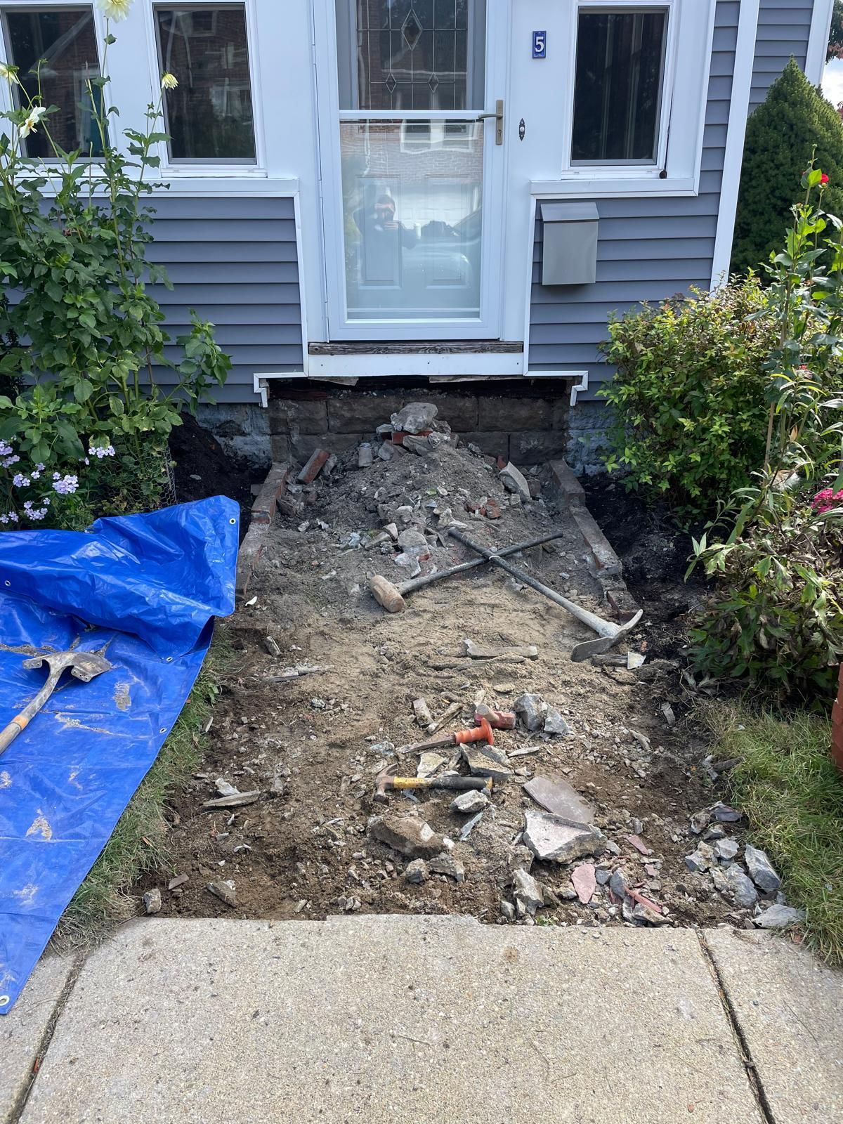 Front entrance construction. Ripped-up concrete pathway in front of a house, debris scattered. Blue tarp on left.