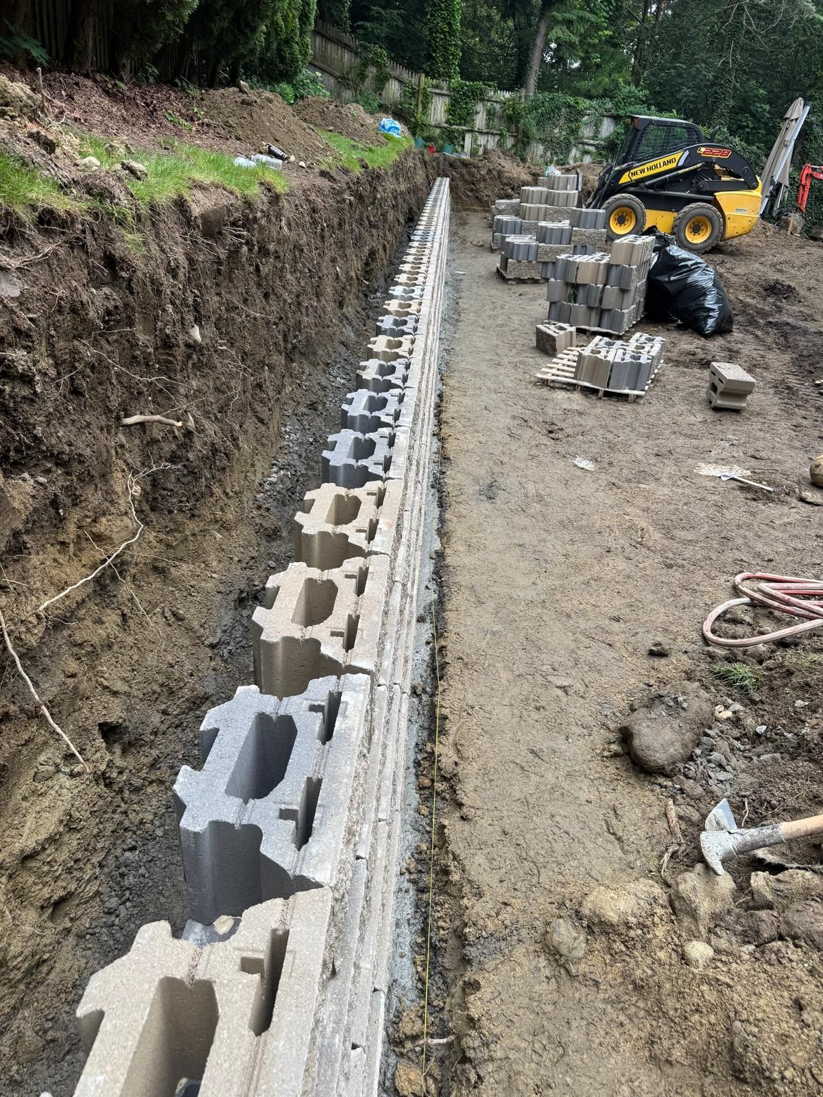 Construction of a retaining wall using concrete blocks in a trench. A small excavator sits in the background.