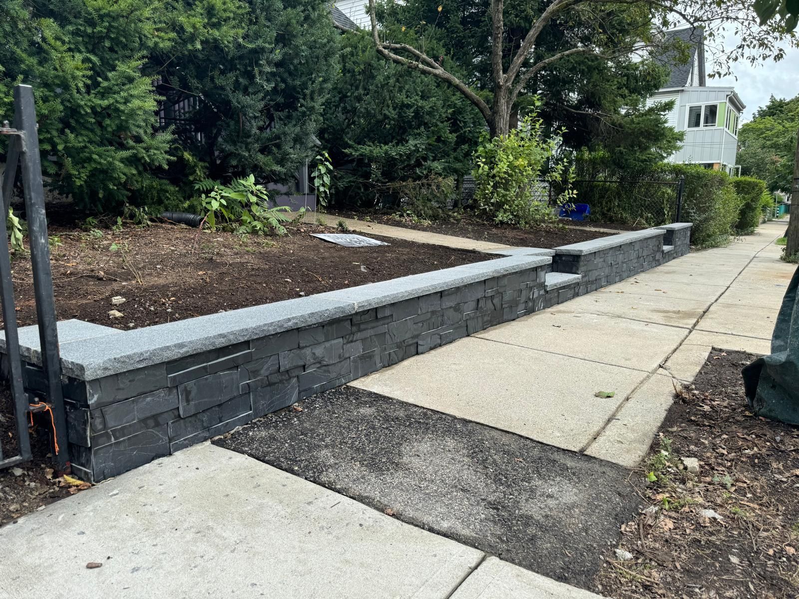 Black stone retaining wall next to a sidewalk and a garden bed.