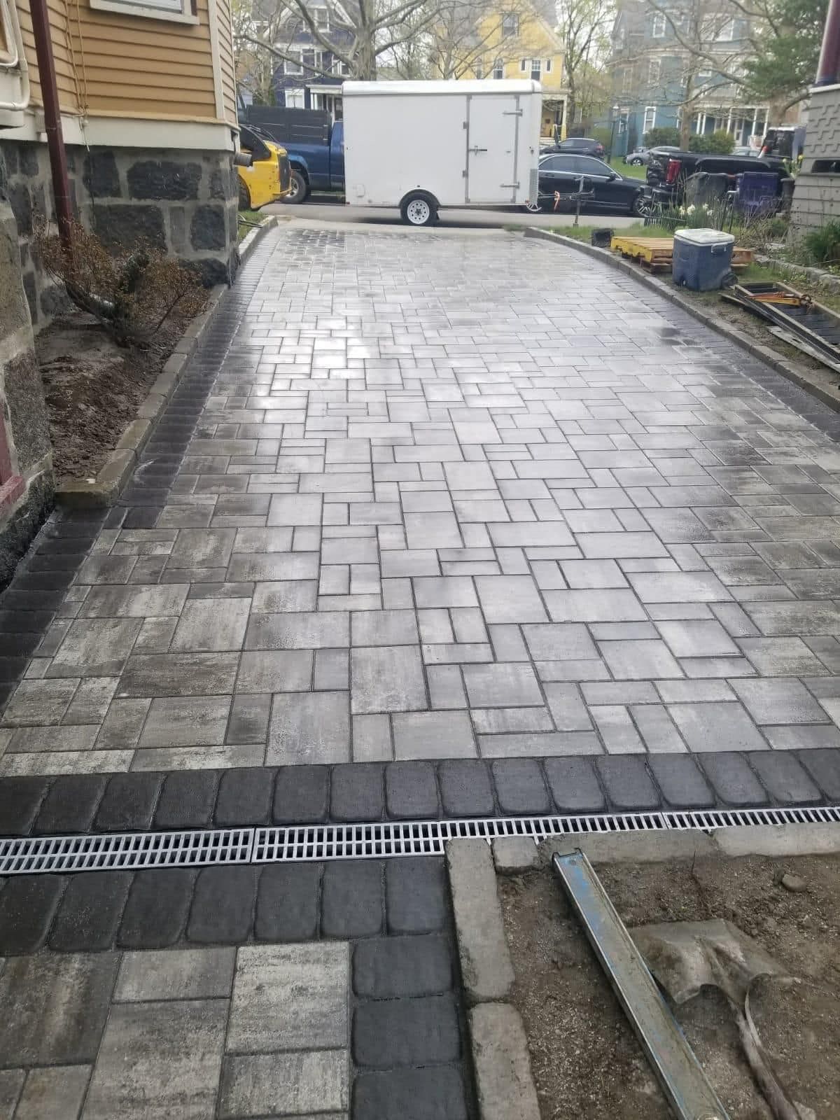 Paver driveway with integrated drainage, beside a house. Trailer and parked vehicles in the background.
