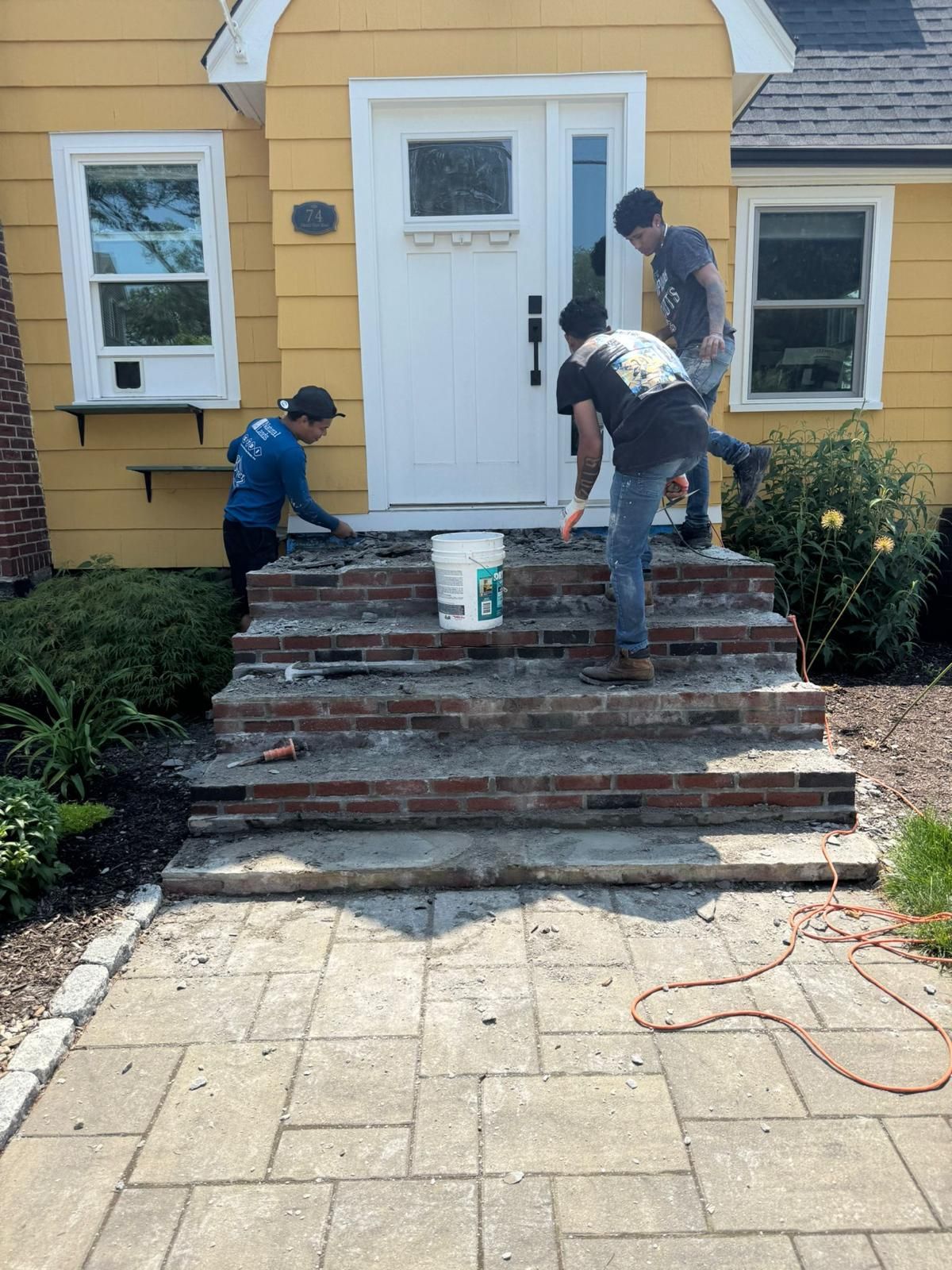 Workers rebuilding brick steps in front of a yellow house with a white door.