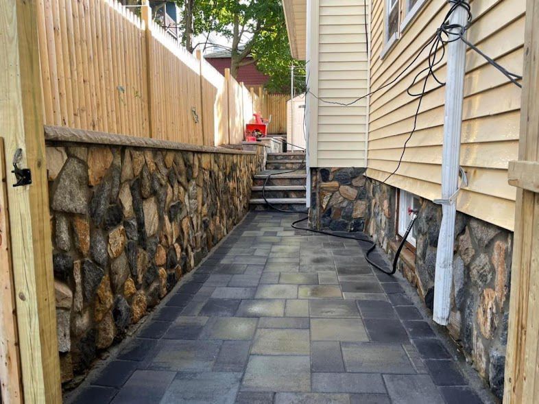 Stone-lined pathway between a wooden fence and a yellow house, with gray pavers and stairs leading to a backyard.