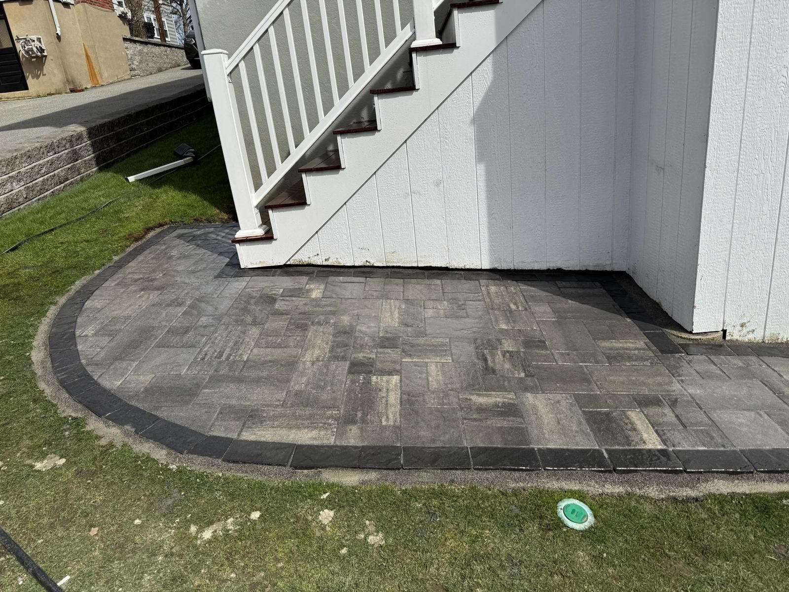 Stone patio with dark border next to white stairs and siding, surrounded by grass.