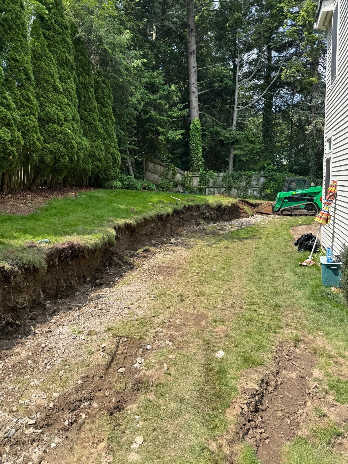 A trench dug in a backyard, next to a house and a lawn. A green machine is visible in the distance.