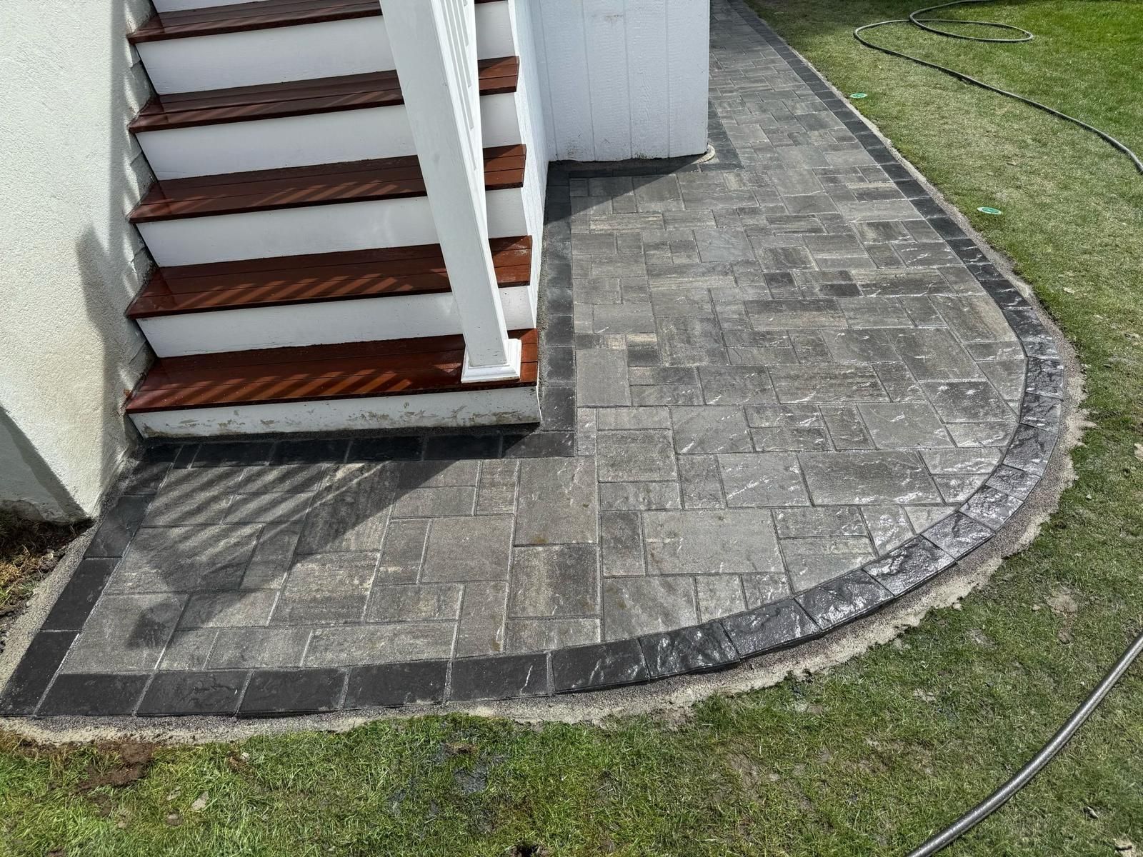Stone patio with curved border adjacent to wooden stairs leading up to a white building.