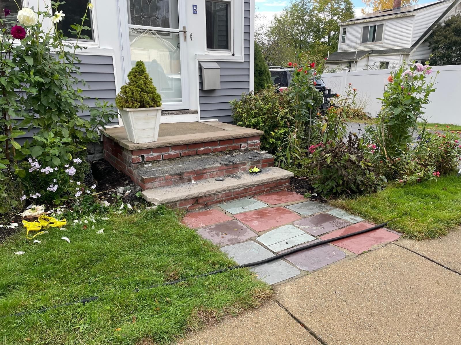 House entrance with brick steps and a paved walkway. Green shrubs and lawn surround.