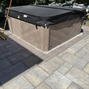 Hot tub with a black cover on a patio with decorative pavers.