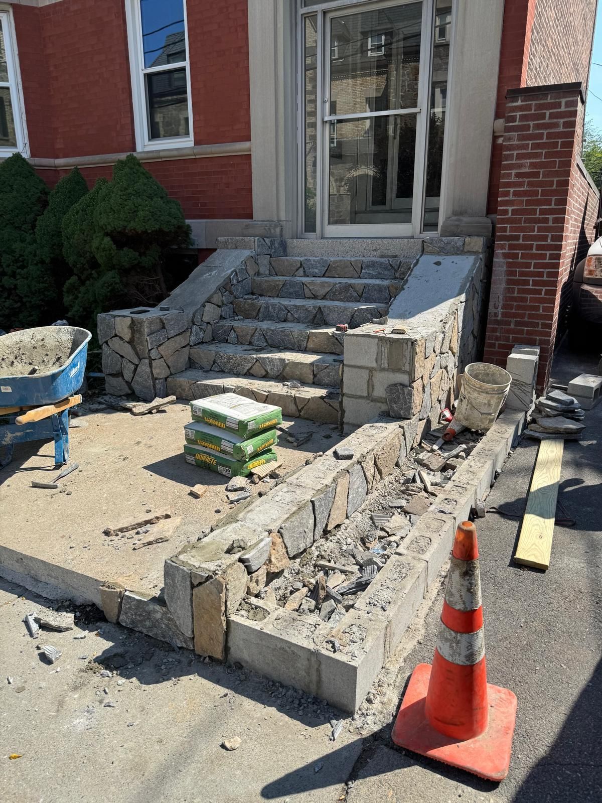 Exterior stairs under construction; stone and concrete steps lead to a glass door. A wheelbarrow, materials, and traffic cone are present.