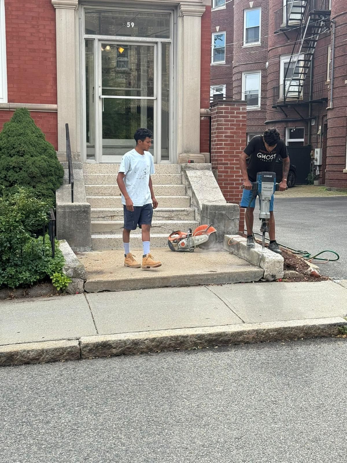 Two people work on concrete steps outside a brick building. One uses a jackhammer.