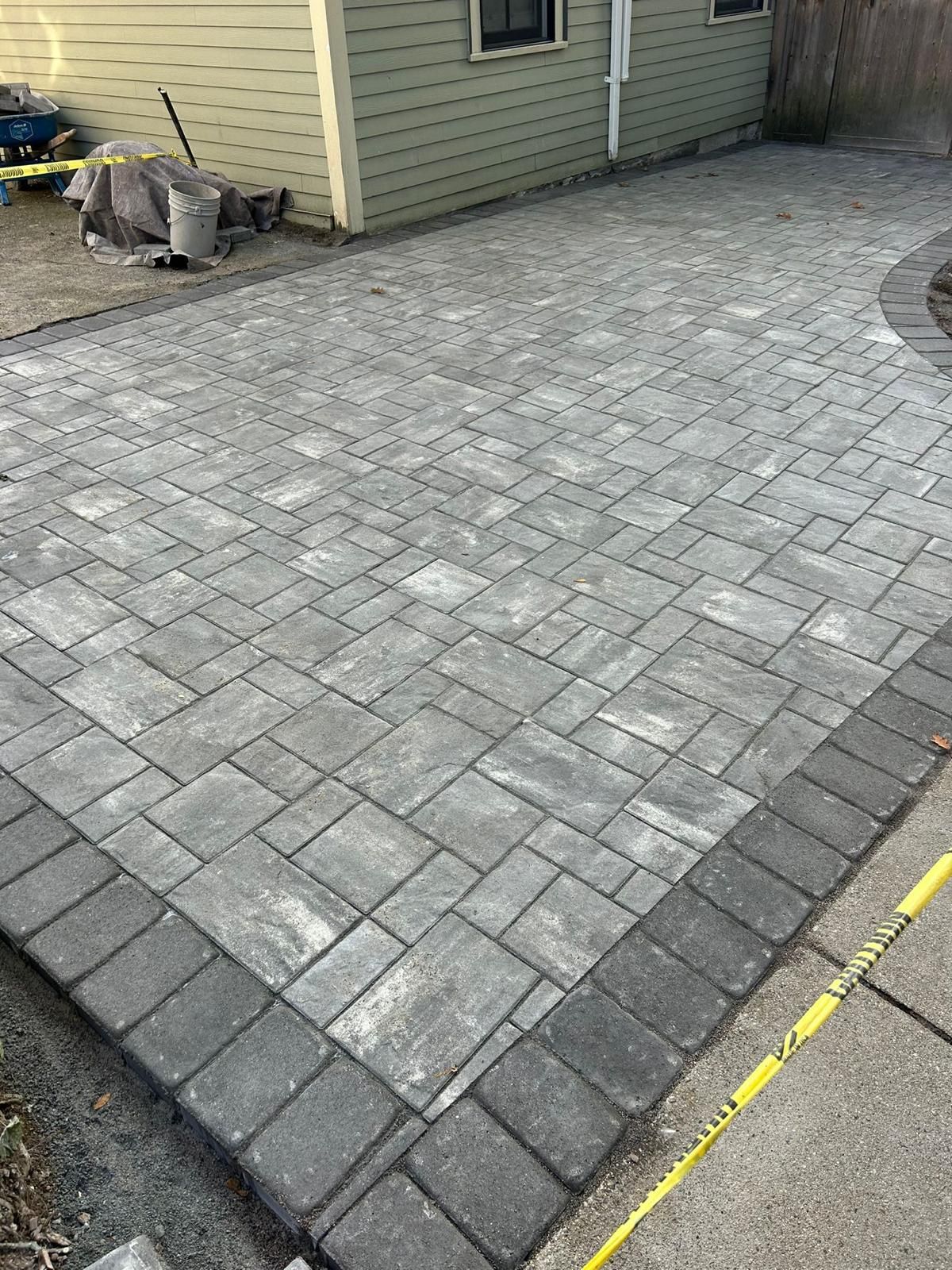 A gray brick patio with a dark border next to a beige building, with caution tape visible.