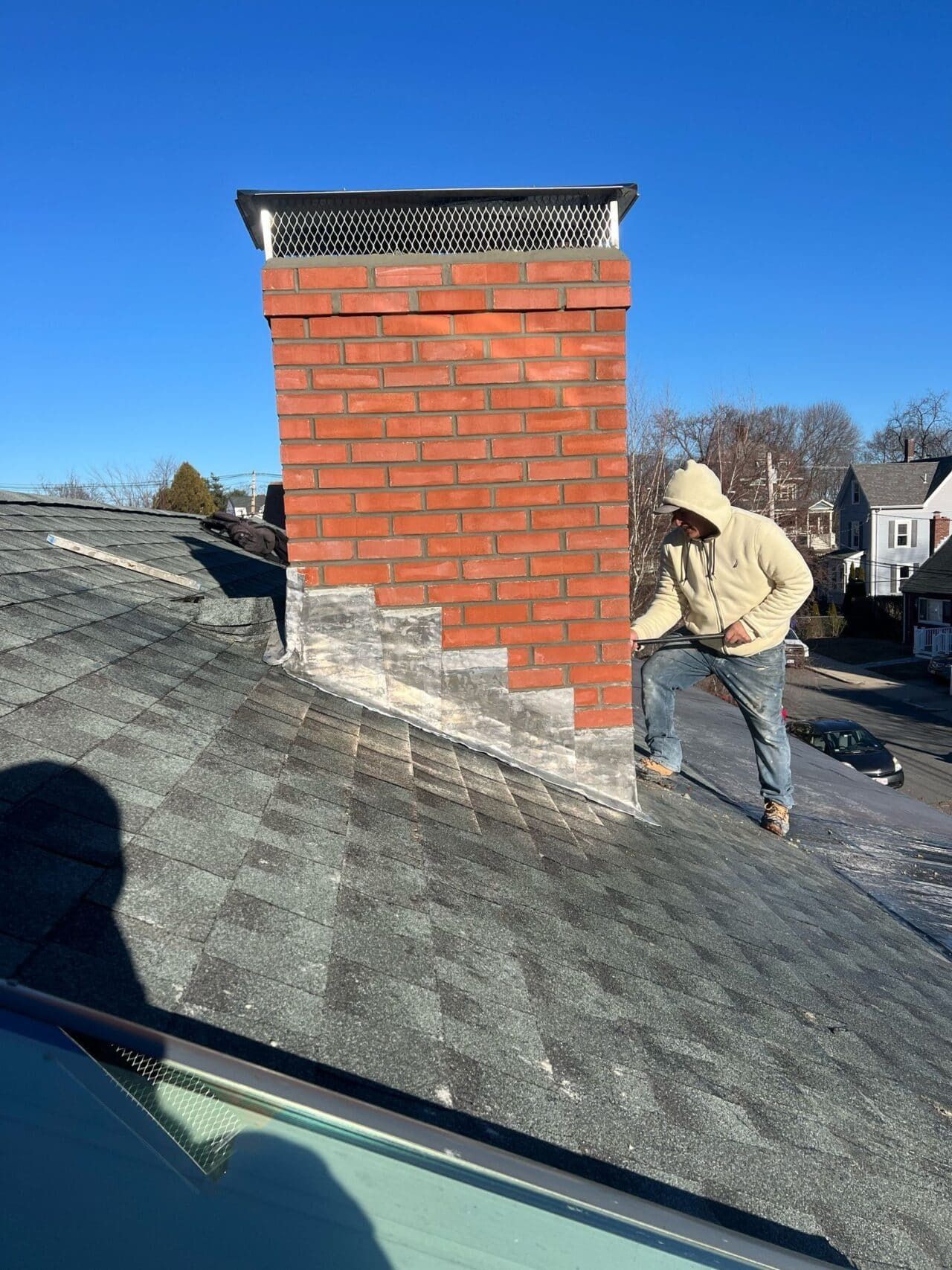 Person on a roof inspecting a brick chimney. Bright blue sky. Roof is covered in dark shingles.