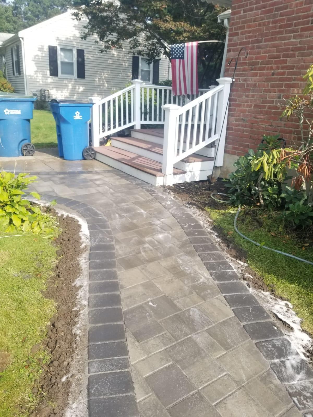 Brick walkway leading to steps with white railing and American flag. Blue trash cans visible.