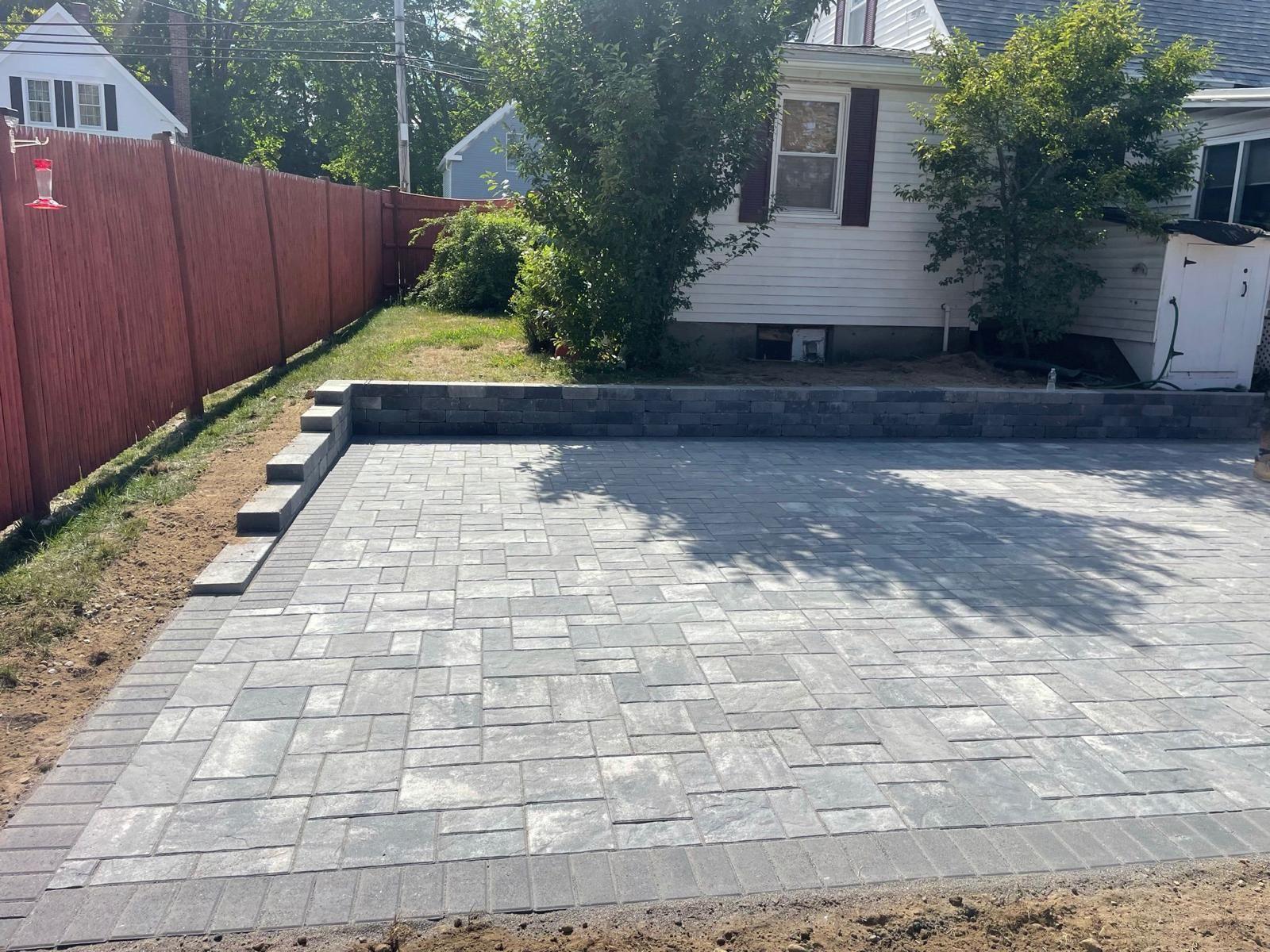 Gray paver patio with a brick border, next to a red fence and small white house.