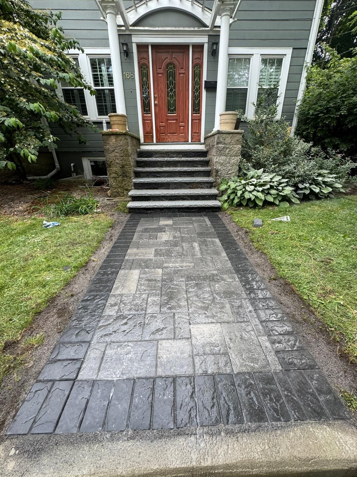 Paver walkway leading to a house with red door and stone steps. Black brick border, green lawn.