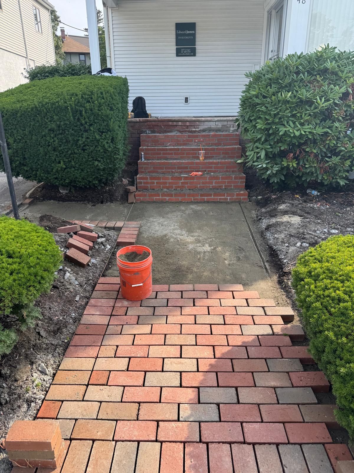 Brick pathway being constructed, leading to brick steps and a white house. Orange bucket visible.
