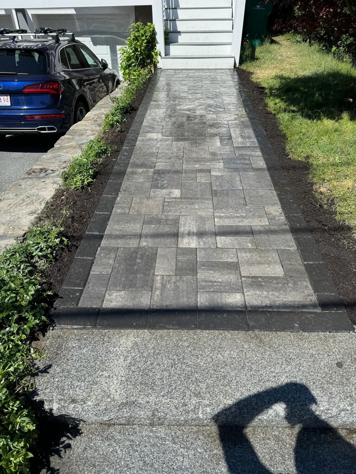 Brick walkway with dark border leading to white house steps. Cars on the left, grass and plants on both sides.