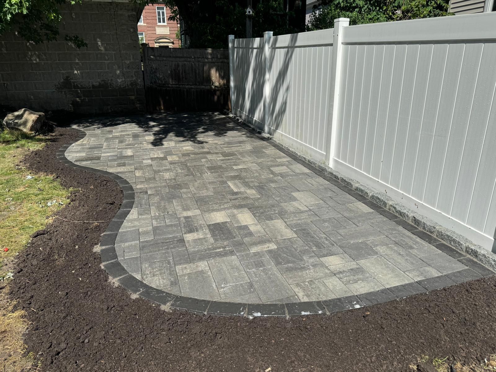 A paved patio bordered by a dark border and a white fence, surrounded by dirt and grass.