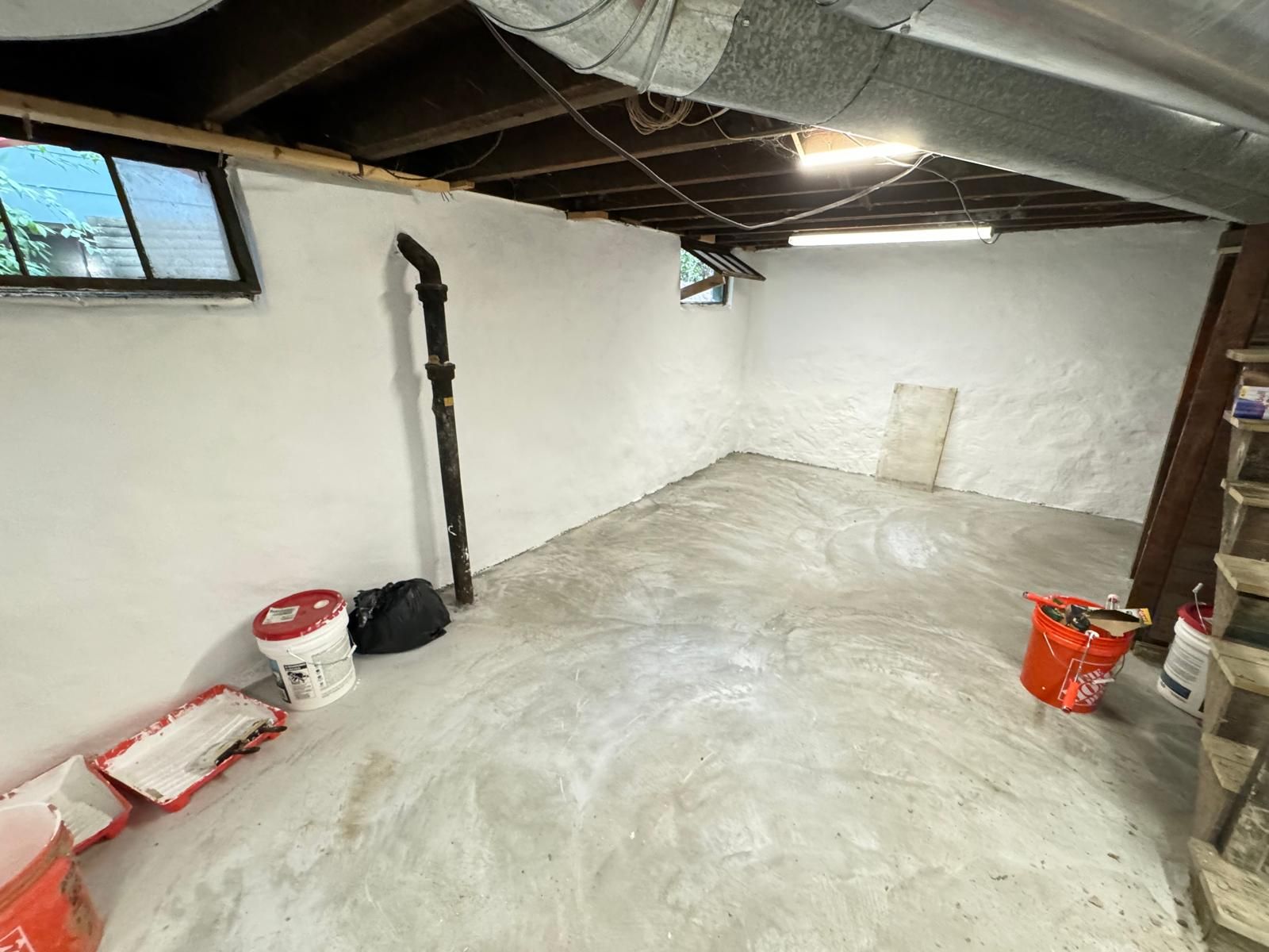 White-painted basement with concrete floor, tools and supplies scattered. Window and pipe visible.