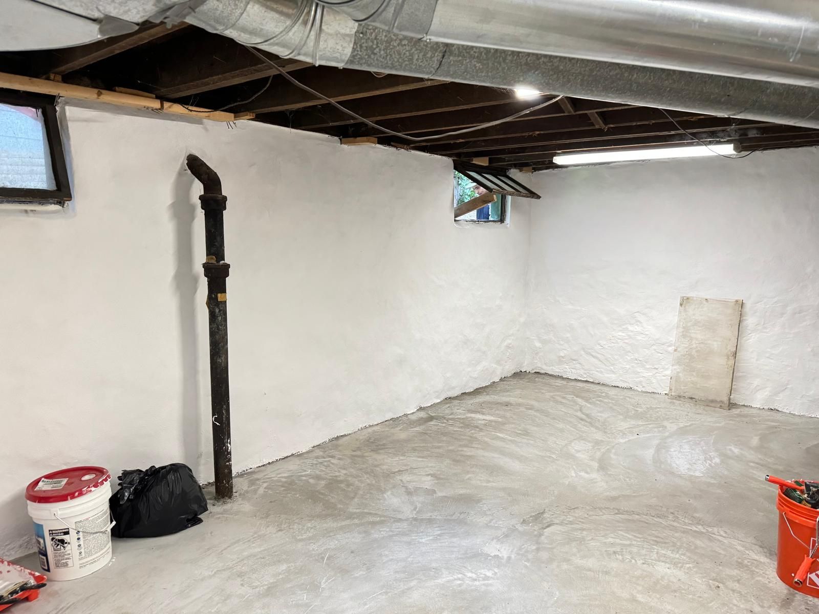 Basement with white-painted walls, concrete floor, pipes, buckets, and a small window.