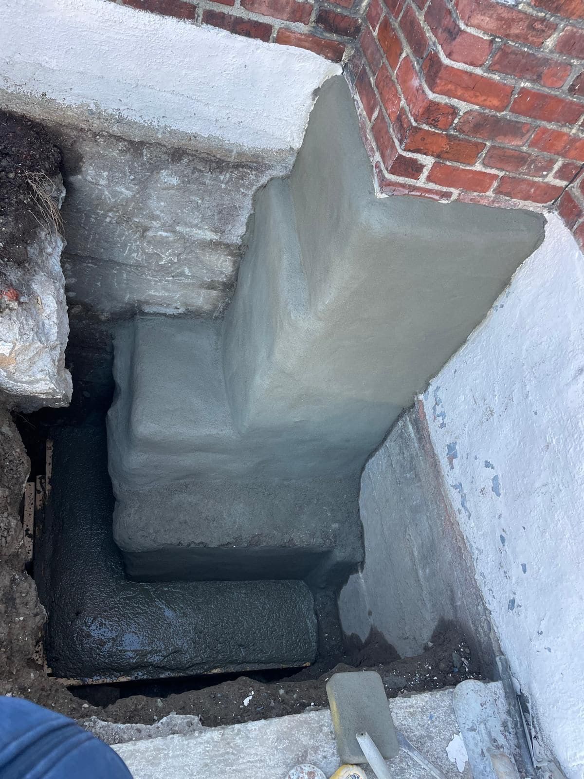 Newly plastered foundation repair next to brick chimney. Light gray cement, excavated dirt visible.