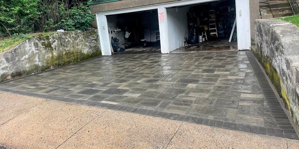 Wet paved driveway leading to two garage doors; concrete retaining walls with vegetation.