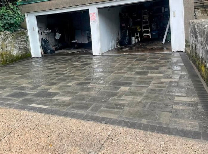 Wet paved driveway leading to two garage doors; concrete retaining walls with vegetation.