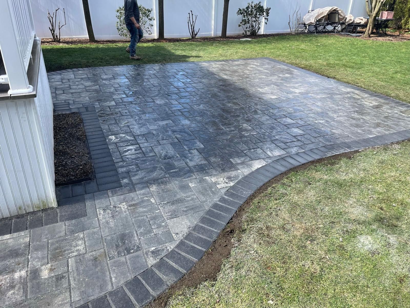A brick patio bordered by dark bricks and a grassy yard. A person is walking in the background.