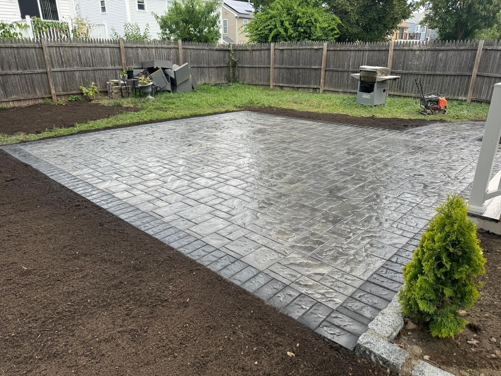 A gray brick patio with a dark border, bordered by grass and dark soil, with a wooden fence.