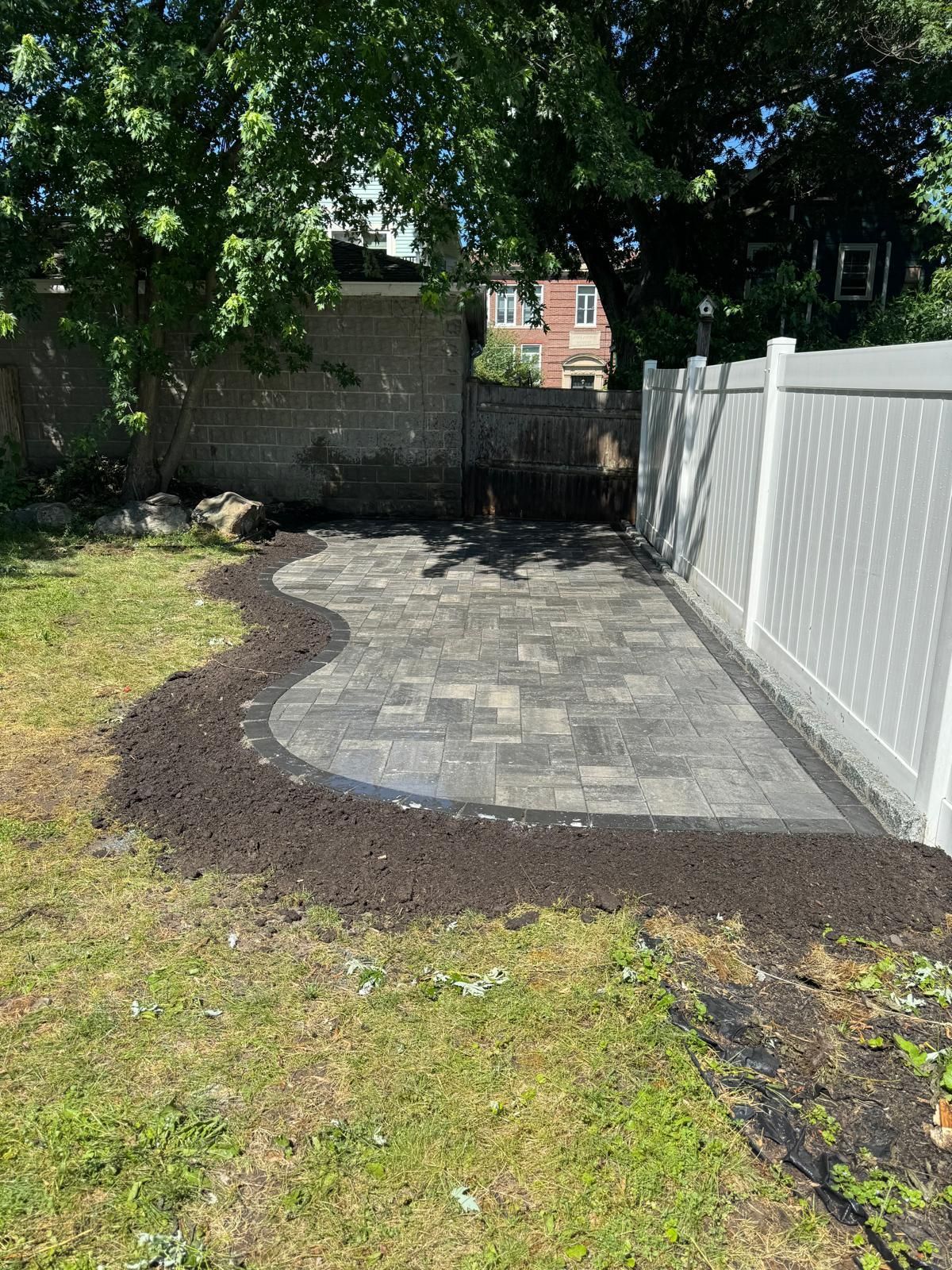Backyard patio with gray pavers and dark soil border, next to a white fence.