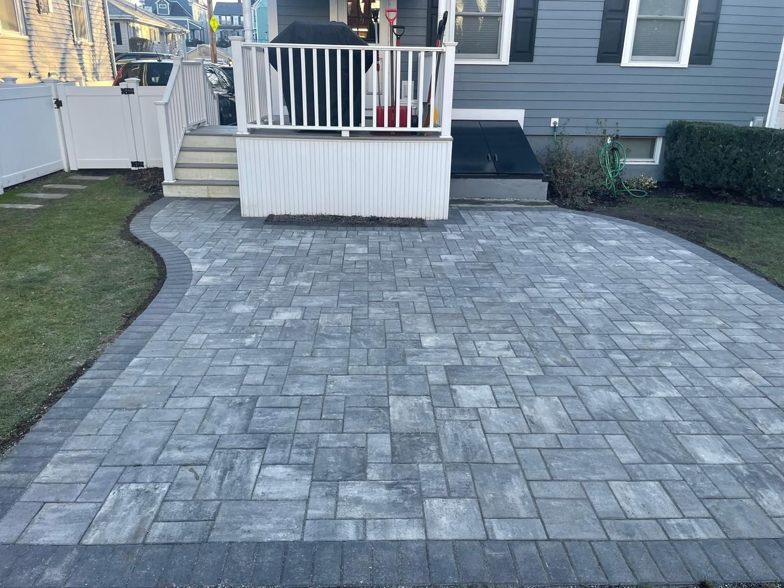 Paver patio with a curved edge, adjacent to a house with a deck and steps, surrounded by grass.