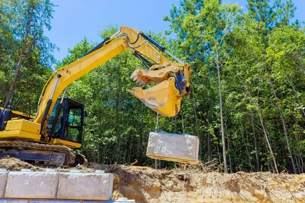 Yellow excavator lifting a large gray concrete block, preparing to place it; outdoors, sunny.
