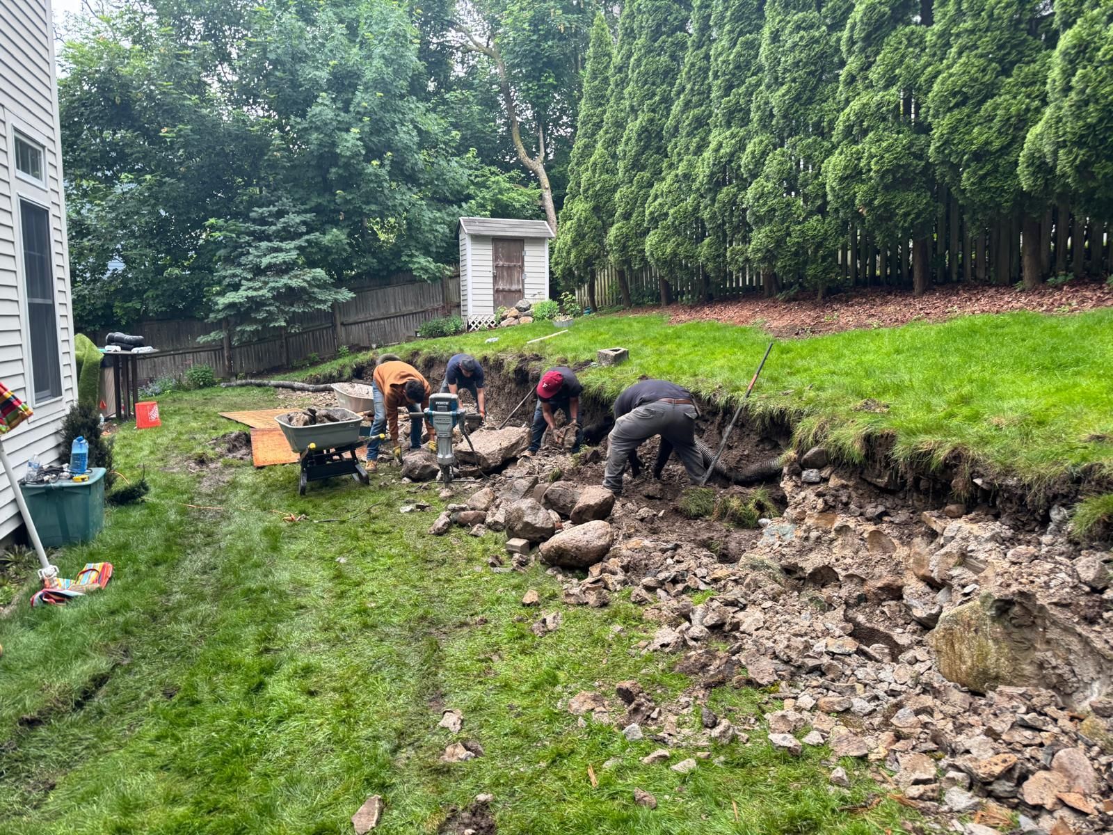 Workers constructing a retaining wall in a backyard, digging in the dirt. Green grass and trees surround them.