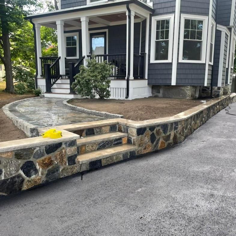 Stone retaining wall with steps leading to a house with a porch and dark siding. Asphalt driveway.