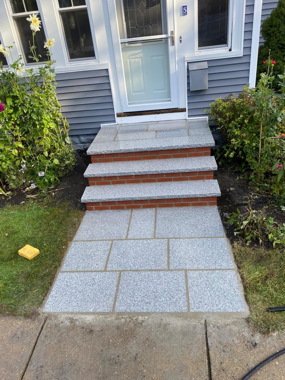 Granite steps and walkway leading to a house entrance. Brick accents on steps, surrounded by greenery.