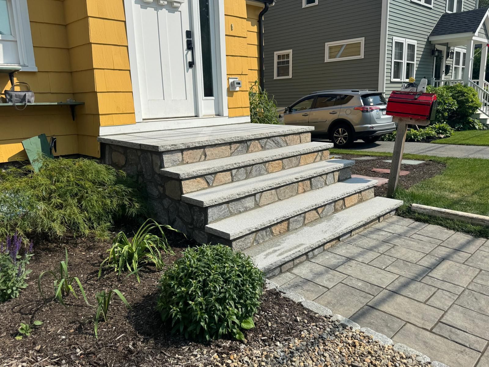Stone steps leading up to a yellow house with a white door. A red mailbox is on the right.