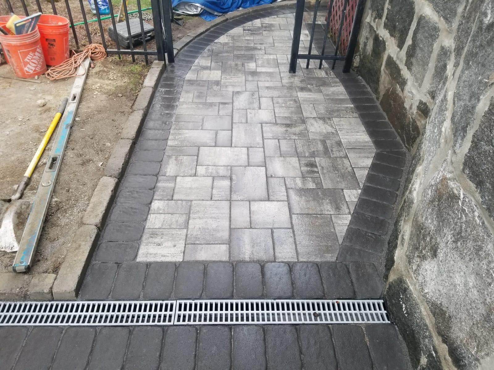Brick pathway leading to a gate, with a gray and dark brick pattern and a drainage grate.