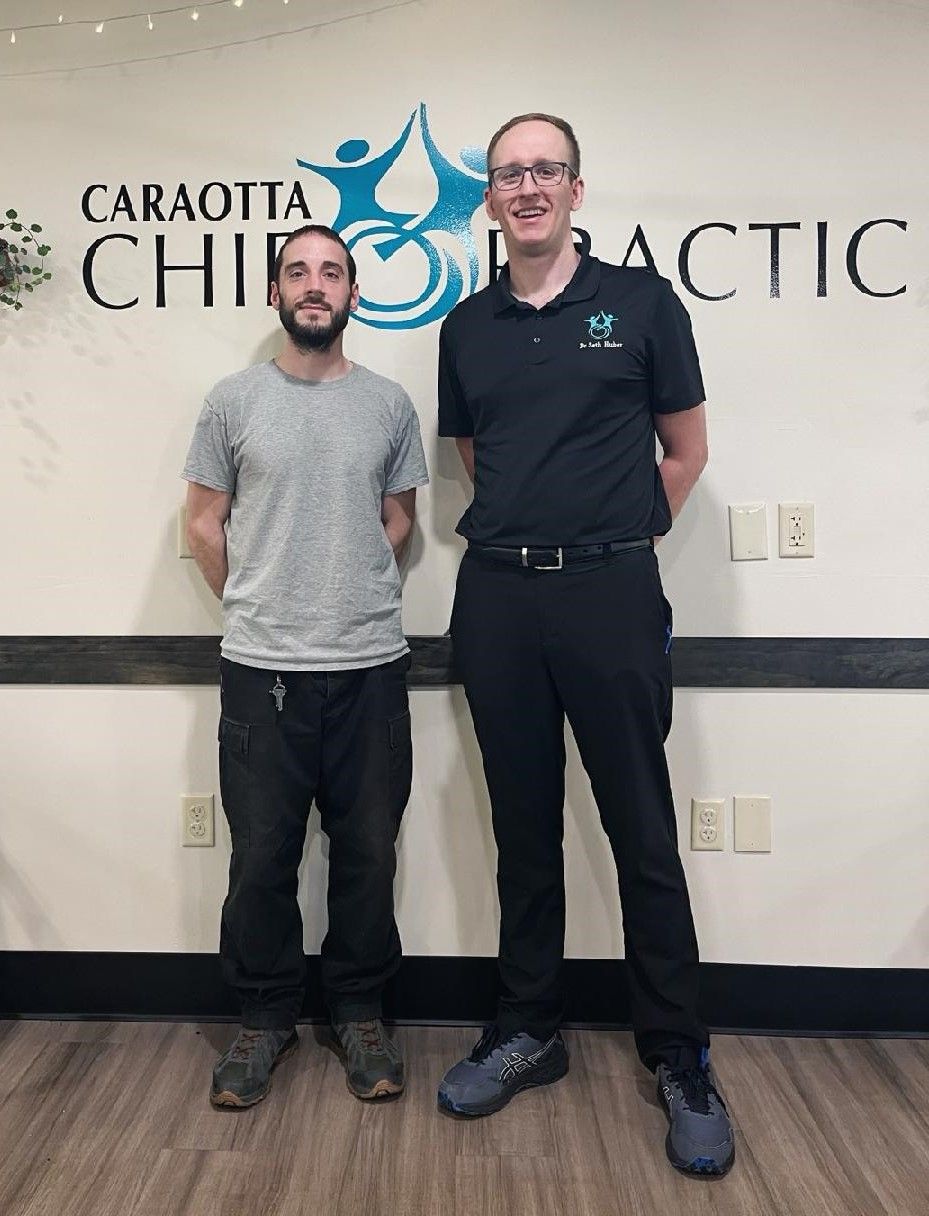 Two people standing in a chiropractic office, smiling in front of a wall sign.