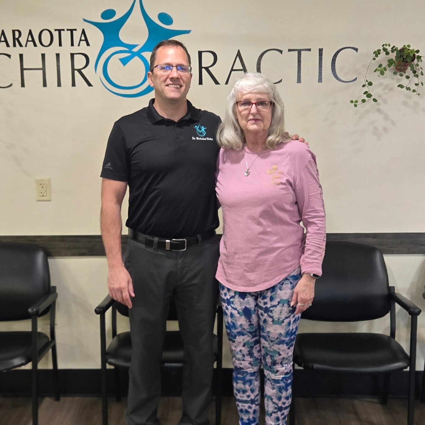 Two people standing in a chiropractic office, smiling in front of a wall sign.