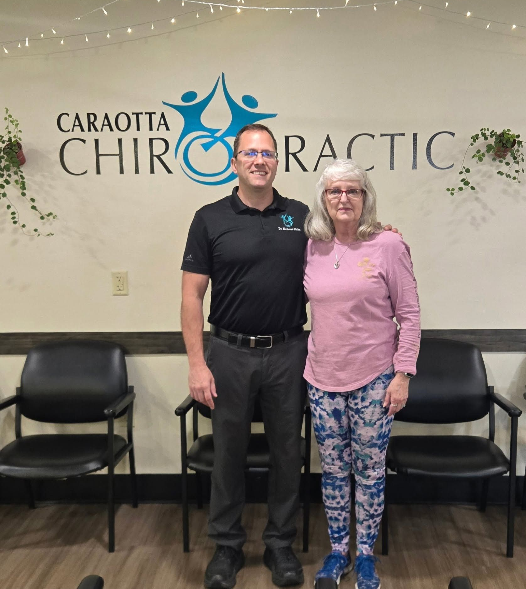 Two people standing in front of a chiropractic office sign, smiling, with chairs and wall decor behind them.