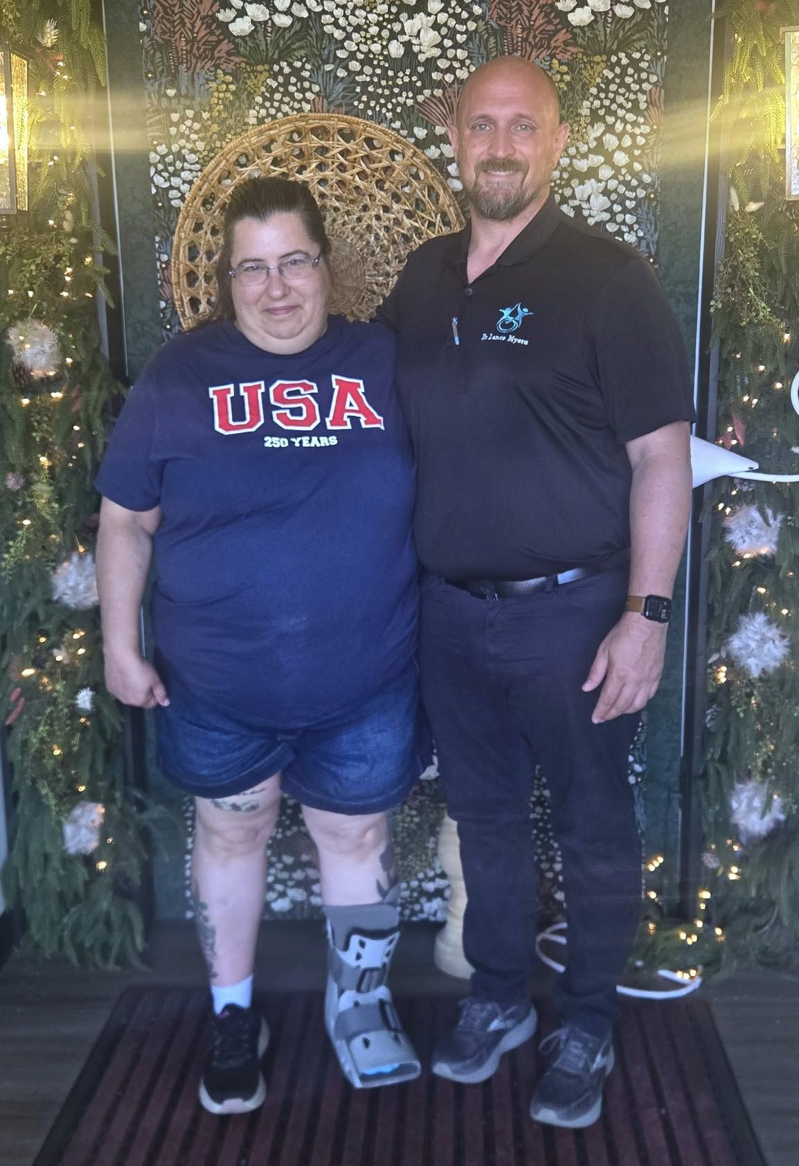 Two people standing together indoors, one in a USA shirt and leg boot, in front of a decorated backdrop