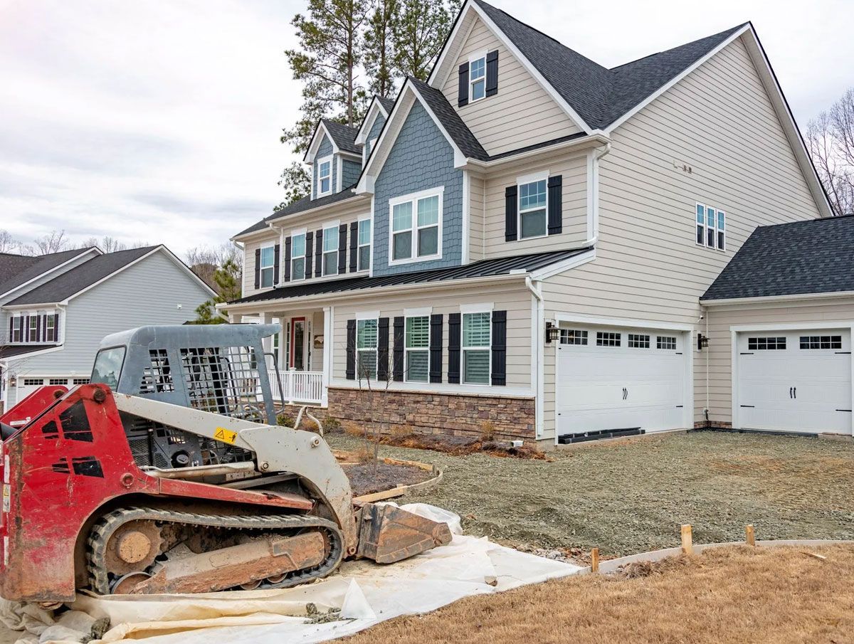 A red skid steer loader sits on a construction site in front of a newly built, multi-story home with beige and blue siding.